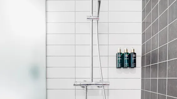 Skinnende krombruserhoved fastgjort mod hvide fliser, ved siden af tre grønne flasker mærket "Hand wash" og "Shower gel," monteret på en grå flisevæg.