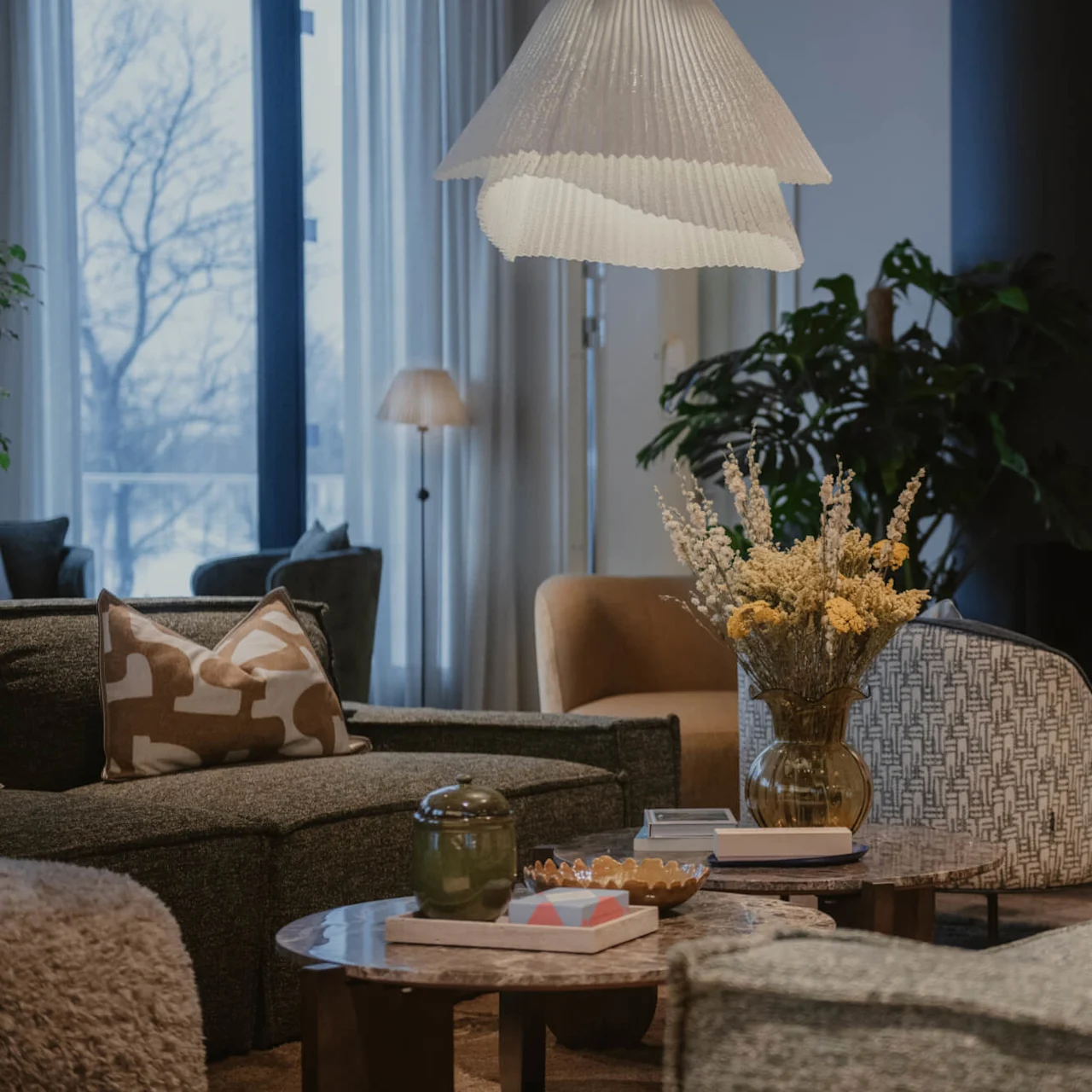 Ett vardagsrum med en hängande lampa lyser upp en mysig miljö. Soffor och fåtöljer omger ett soffbord med böcker och blommor i en vas. Stora fönster släpper in naturligt ljus.