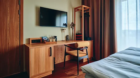 Et moderne hotellrom med en komfortabel seng, et treskrivebord med TV og en stol, badet i naturlig lys fra et vindu.