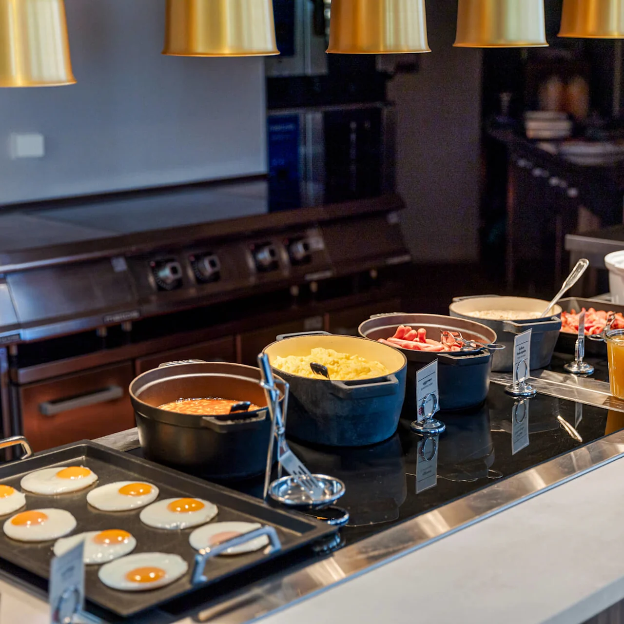 A breakfast buffet displays various foods on a counter with serving utensils in a well-lit kitchen area.