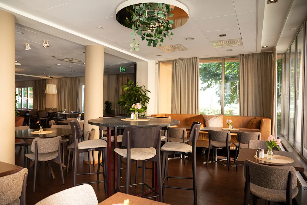 En modern och inbjudande hotellounge eller restaurang med olika bord och sittplatser, inklusive en lång soffa vid stora fönster. EXIT-skylt synlig.