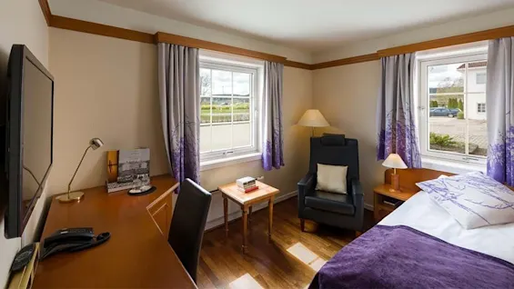 Et rent hotelværelse har en seng med lilla detaljer, et skrivebord med lampe og telefon, og en lænestol ved to vinduer der viser en parkeringsplads og marker.
