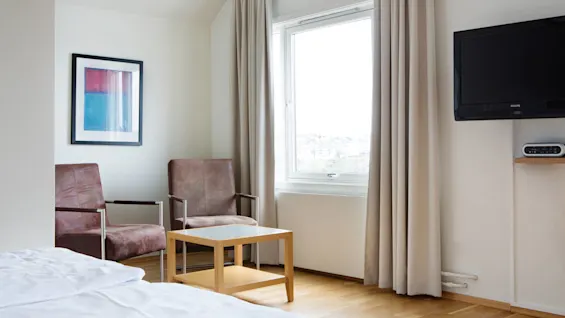 To brune stoler står ved siden av et trebord i et lyst rom. Et vindu med gardiner har utsikt over bylandskapet. En veggmontert TV og abstrakt kunstverk fullfører innredningen.