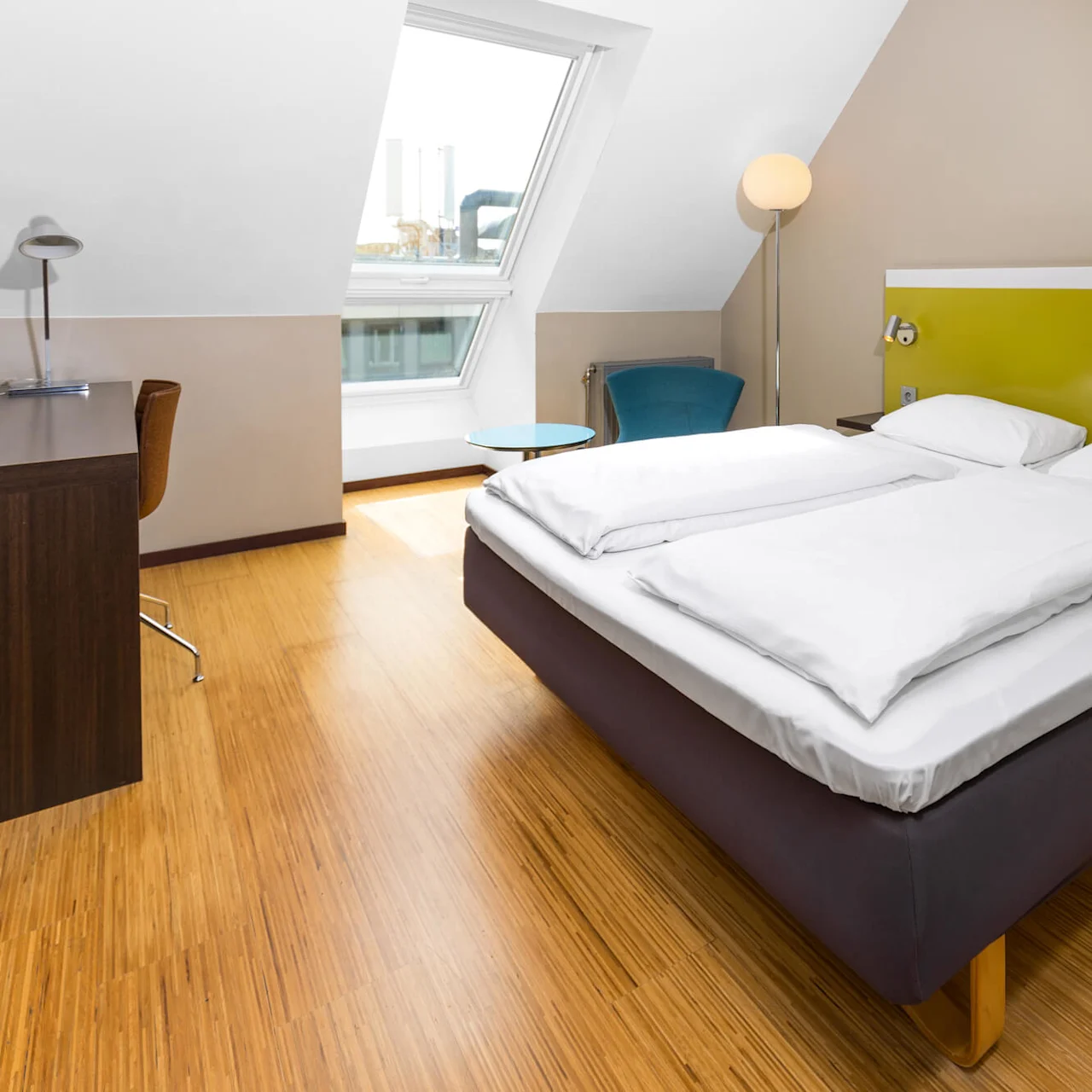 A double bed with white linens sits in a modern, bright room. A wooden desk with a chair is nearby, alongside a window, blue chair, and floor lamp.