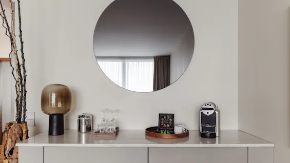 En stilren bänk i ett rum med kaffemaskin, glas och en dekorativ lampa, med en stor rund spegel som reflekterar ett fönster.