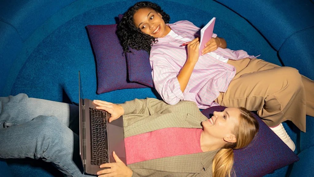 Kaksi henkilöä makaa sinisillä tyynyillä, toinen käyttää kannettavaa tietokonetta ja toinen pitää muistilehtiötä ja kynää rennossa ympäristössä.