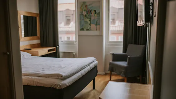 En seng med hvidt sengetøj står i et lille, ryddeligt hotelværelse. En stol og skrivebord vender mod to vinduer med gardiner, med udsigt over hustage. Et maleri hænger over radiatoren.