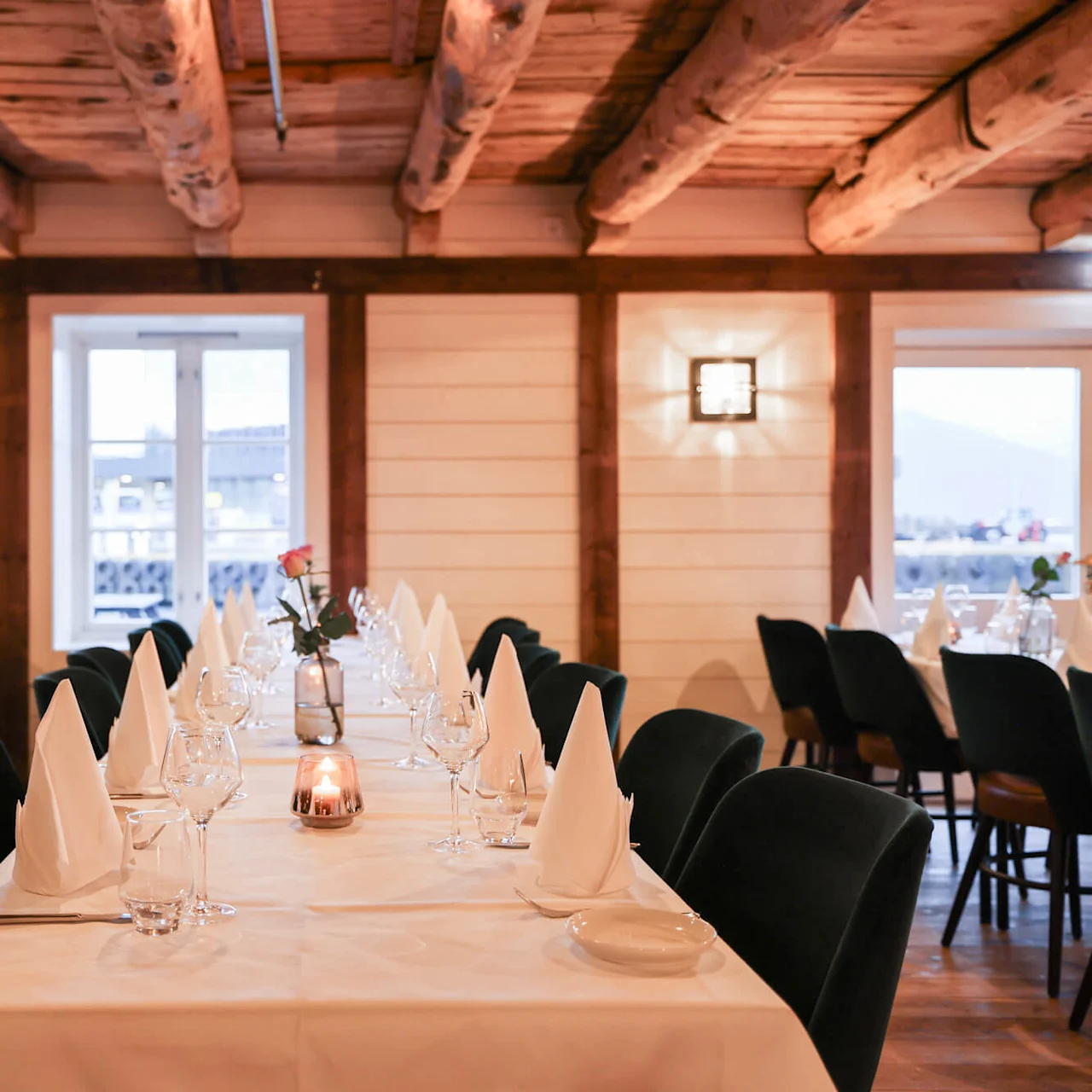 A long dining table, adorned with neatly arranged glasses, napkins, and a single rose in a vase, sits in a rustic wooden room with large windows. 