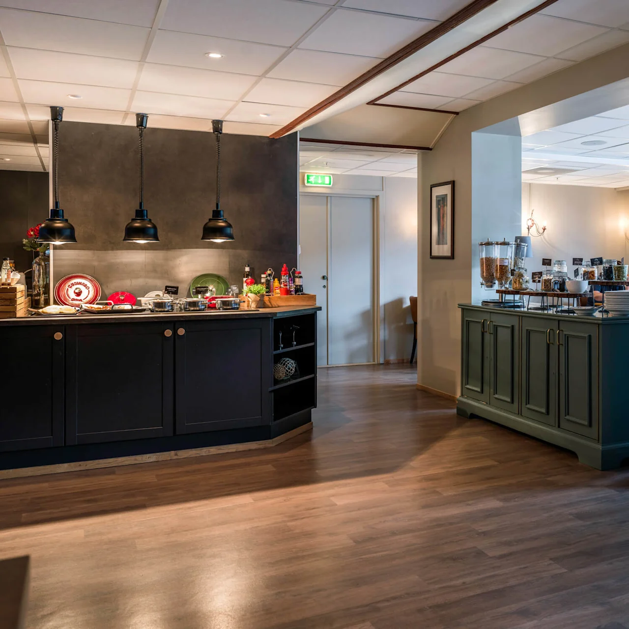 A buffet area features wooden cabinets displaying food items under hanging lights; cereals and bowls are arranged on a sideboard nearby, within a spacious dining room setting.