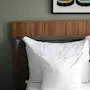 White pillows rest against a wooden headboard in a neatly made bed, beside a black lamp. Above, a colorful abstract artwork decorates the gray wall.