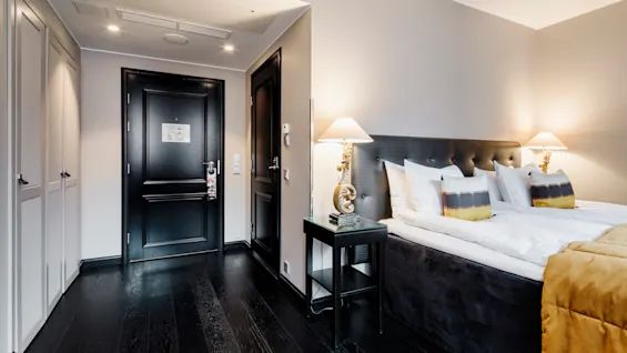 En pent sengeredning med hvite puter og mørk sengegavl står ved siden av to stilige lamper på et elegant hotellrom med mørkt tregulv og lyse vegger.