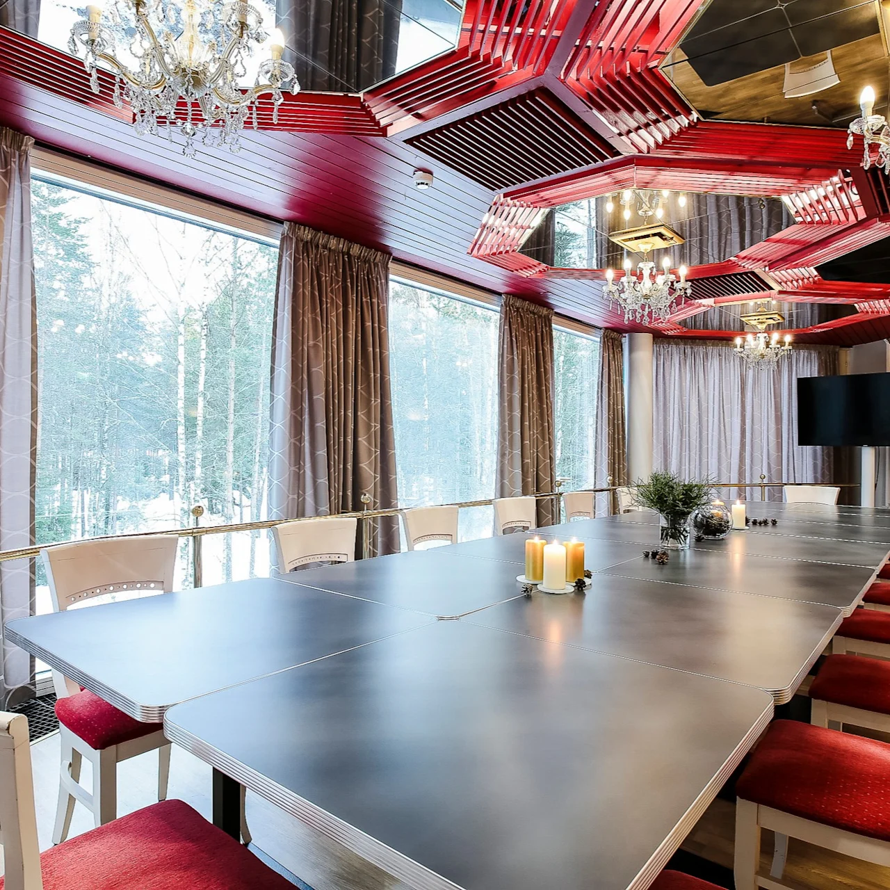 A long conference table with candles and decorations is surrounded by red chairs, located in a room with large windows, patterned curtains, and chandelier lighting. A TV is mounted on the wall.