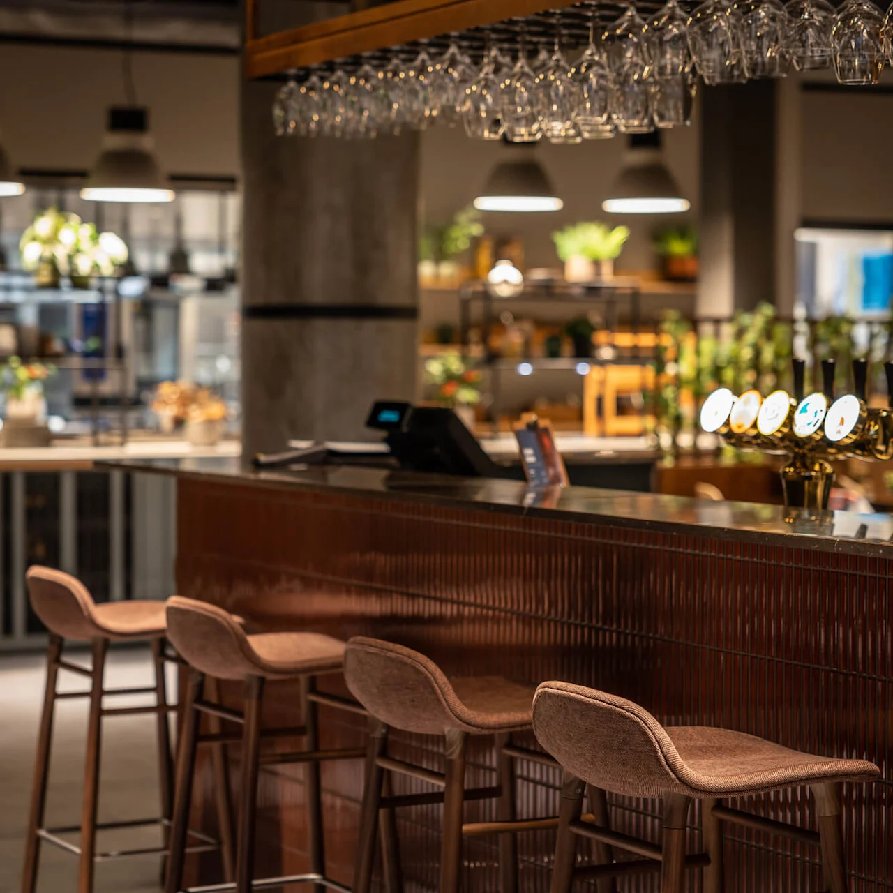 Bardisk med høje stole står tom, omgivet af hængende glas og oplyste ølhaner; hyggelig, varmt oplyst restaurant- eller barindretning med potteplanter og sløret baggrund.