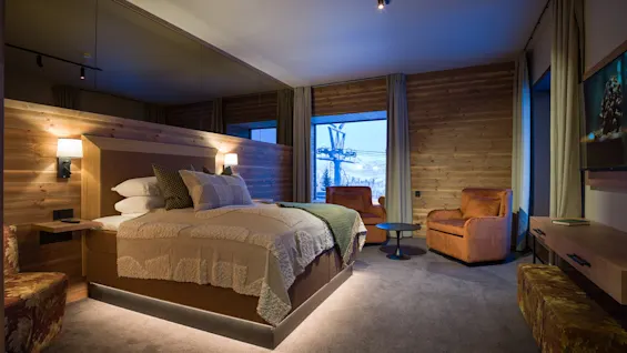 A cozy bedroom features a large bed with patterned bedding, two orange armchairs, and wooden walls. Soft lighting creates a warm atmosphere, and a mountain view is visible outside the window.