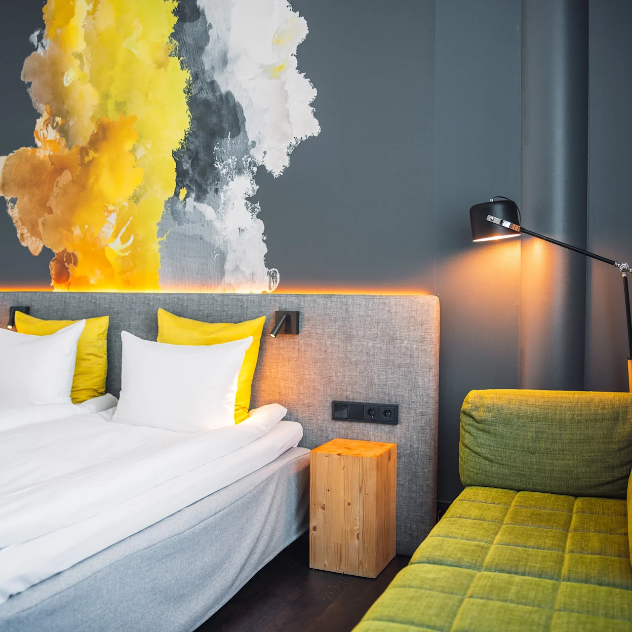 A modern hotel room features a neatly made bed with white and yellow pillows, a vibrant abstract wall painting, a green sofa, and a wall-mounted reading lamp, creating a cozy ambiance.