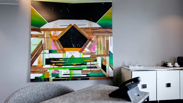 A colorful abstract painting hangs on a gray office wall above a modern stone desk with a telephone. A sideboard with tea set sits in the background.