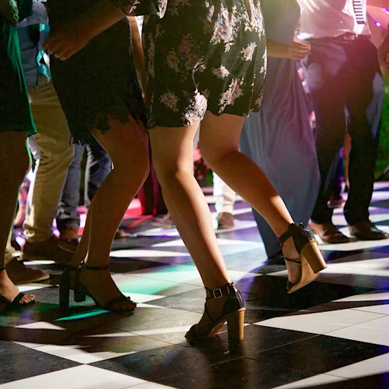 Nærbilde av folk som danser i høye hæler og festantrekk på et sjakkrutete dansegulv med fargerik belysning.