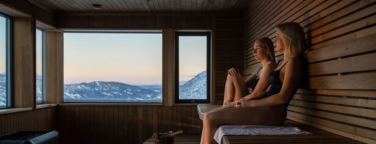 Two women sit relaxing in a wooden sauna, gazing out at a snowy mountain landscape through large windows, creating a serene and warm atmosphere.