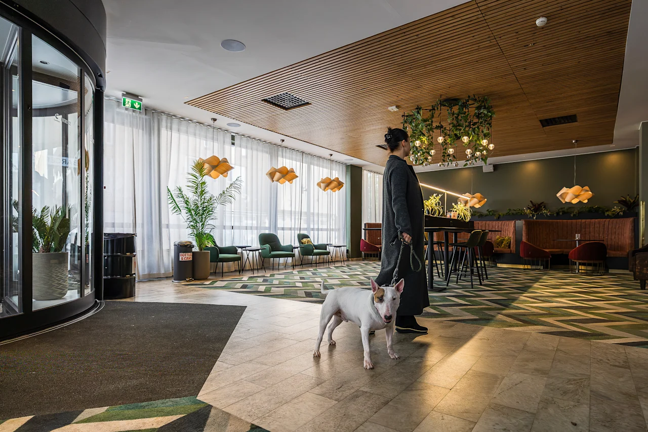 En person spaserer gjennom en elegant hotellobby med en hund, omgitt av frodige planter og moderne sitteplasser. Varme pendellamper skaper en koselig atmosfære, med geometriske mønstre på gulvet.