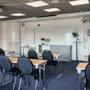 Et klasseværelse med rækker af tomme sorte stole og træborde vendt mod en whiteboard. Loftlamper oplyser rummet. Et ur og projektor ses på vægge og loft.