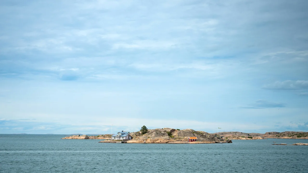 En lille, klippefyldt ø med nogle bygninger står i et roligt hav under en skyet himmel, med fjernt land synligt i horisonten.