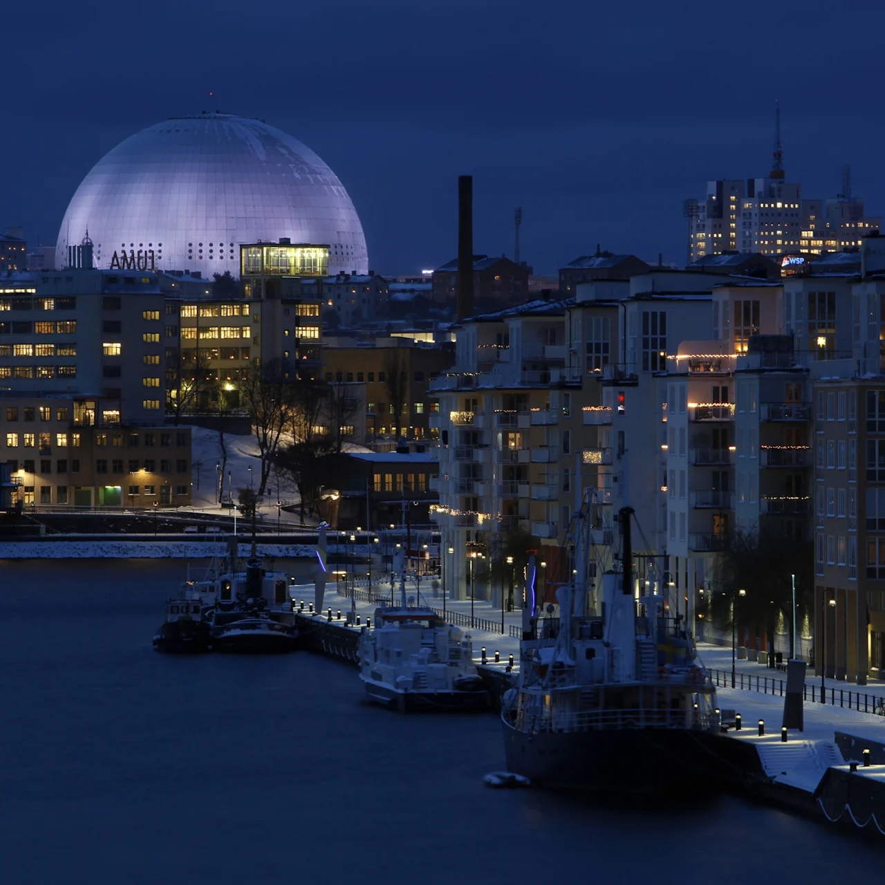 Stadsvy på natten med en stor upplyst kupol, upplysta byggnader och båtar vid en snötäckt kaj. Text: AMOI.
