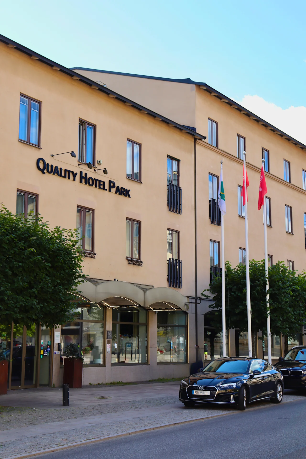 Eksteriør av Quality Hotel Park Södertälje med biler parkert på gaten og flagg som vaier foran bygningen.