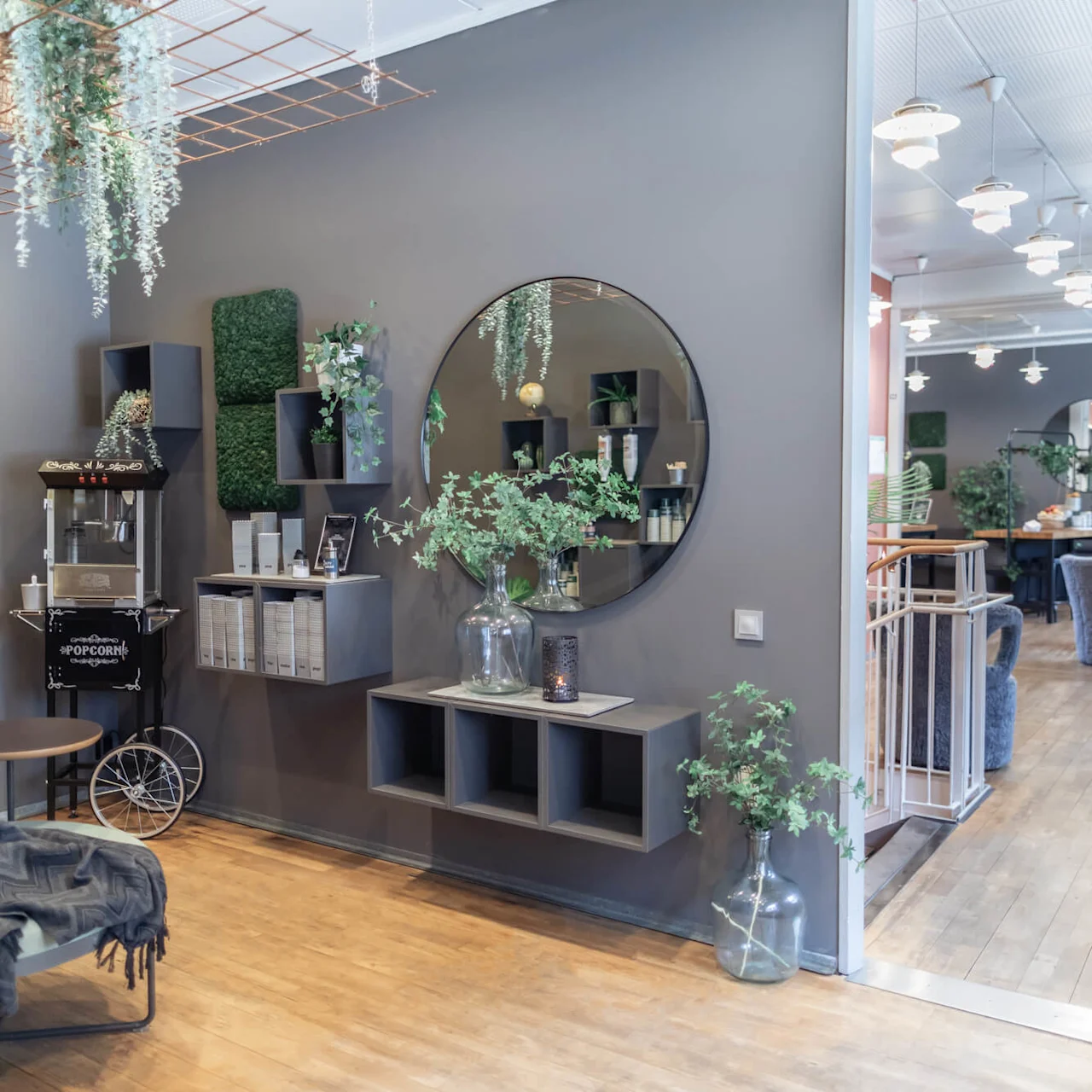 A popcorn machine on wheels stands in a modern lounge with grey walls and green decor, featuring shelves, a large mirror, plants, and contemporary seating.