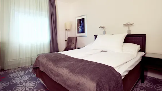 A neatly made bed with white linens in a well-lit hotel room, including a dark upholstered chair and a framed picture on the wall.