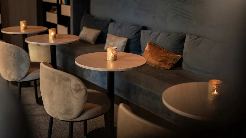 Et hyggeligt, dæmpet loungeområde med små borde, komfortable stole og en lang bænk, hvert bord med et flimrende lys.