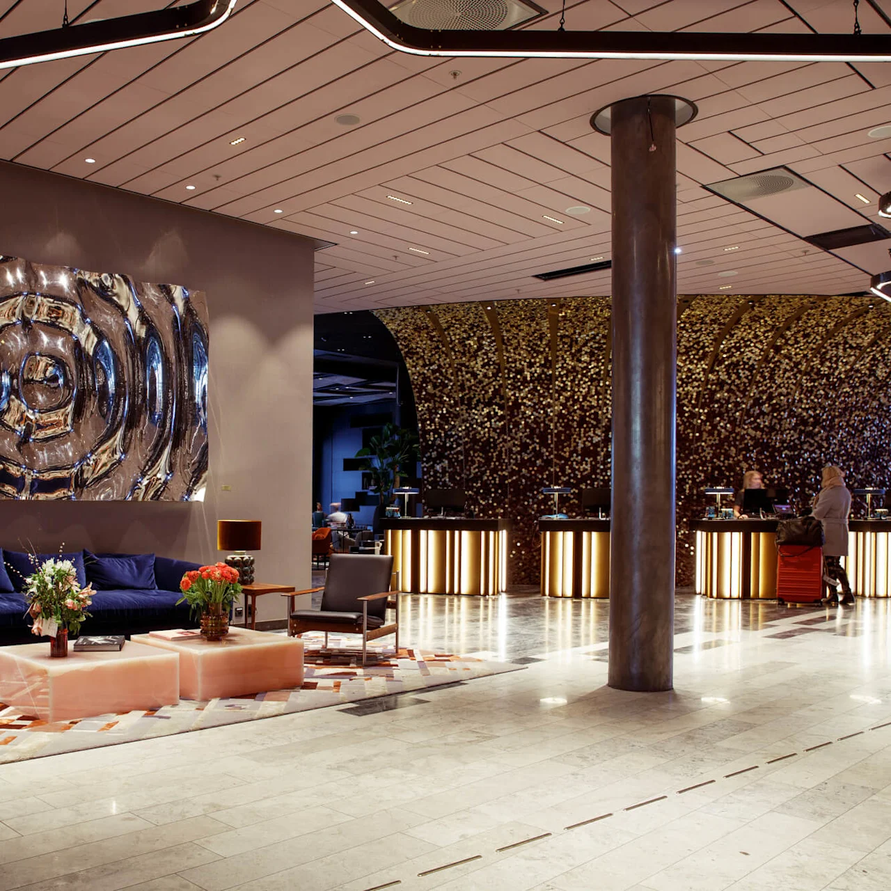 A spacious hotel lobby with modern seating arranged on a patterned rug, abstract art on the wall, and a reception desk featuring illuminated panels adorned with metallic decor.