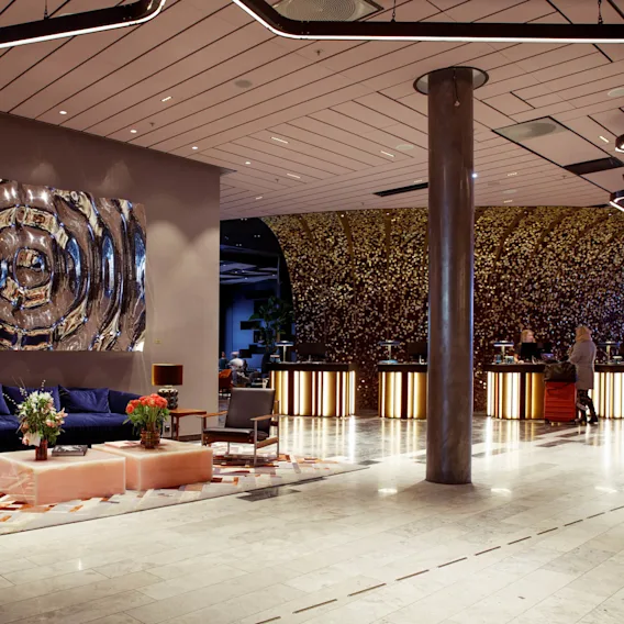A spacious hotel lobby with modern seating arranged on a patterned rug, abstract art on the wall, and a reception desk featuring illuminated panels adorned with metallic decor.