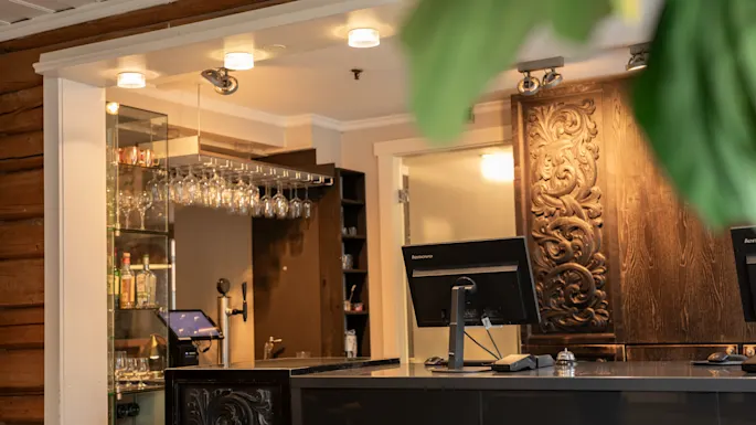 A bar counter supports two computer screens in a warmly lit space. Glass shelves display bottles and glasses. Intricately carved wooden panels adorn the walls.