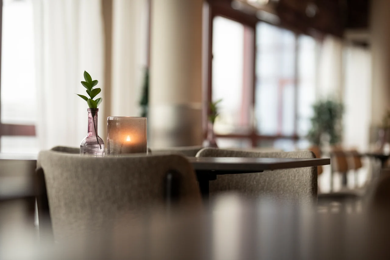 Viihtyisä sisätila, jossa on pöytä, tuoleja, pieni kasvi maljakossa ja sytytetty kynttilä, suuret ikkunat taustalla.