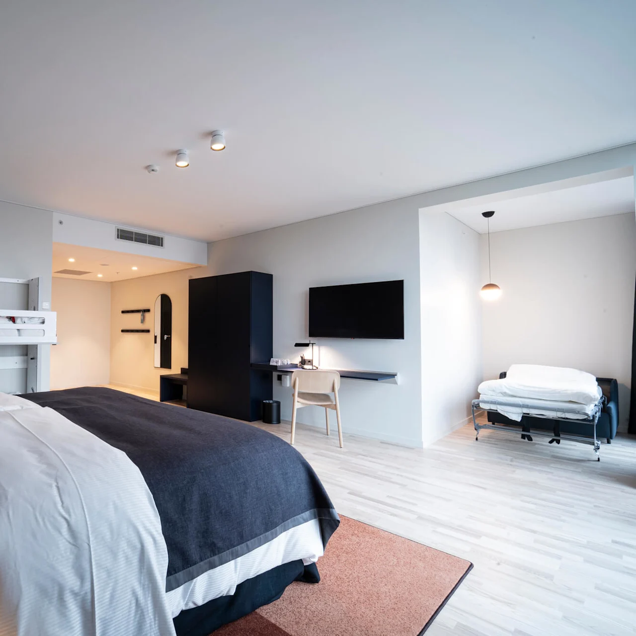 En stor seng med hvidt og mørkt sengetøj er i et rummeligt moderne hotelværelse, med vægmonteret tv, skrivebord, stol og en udtræksseng i en alkove.