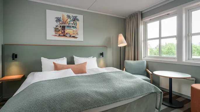 A neatly made bed with green covers is centered in a modern hotel room, flanked by small nightstands. A floor lamp stands nearby, and a window shows greenery outside.