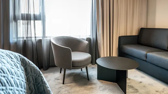 En grå lænestol står overfor et sort rundt sofabord. I nærheden står en mørkegrå sofa mod beige gardiner, der filtrerer lys fra et vindue.
