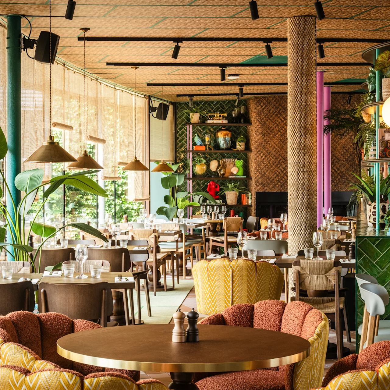 A restaurant with colorful furniture and tropical plants inside, tables set with glasses and cutlery, surrounded by large windows letting in natural light.