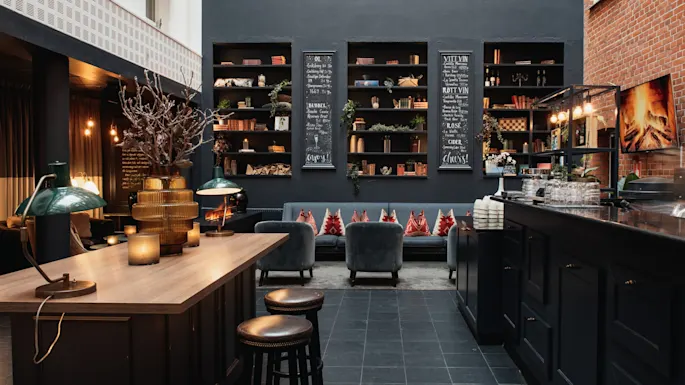 En varm og innbydende lounge med bar, komfortable sitteplasser og bokhyller. Krittavler viser OL, BUBBEL, VITT VIN, ROTT VIN, ROSE, CIDER, cheers, enjoy.