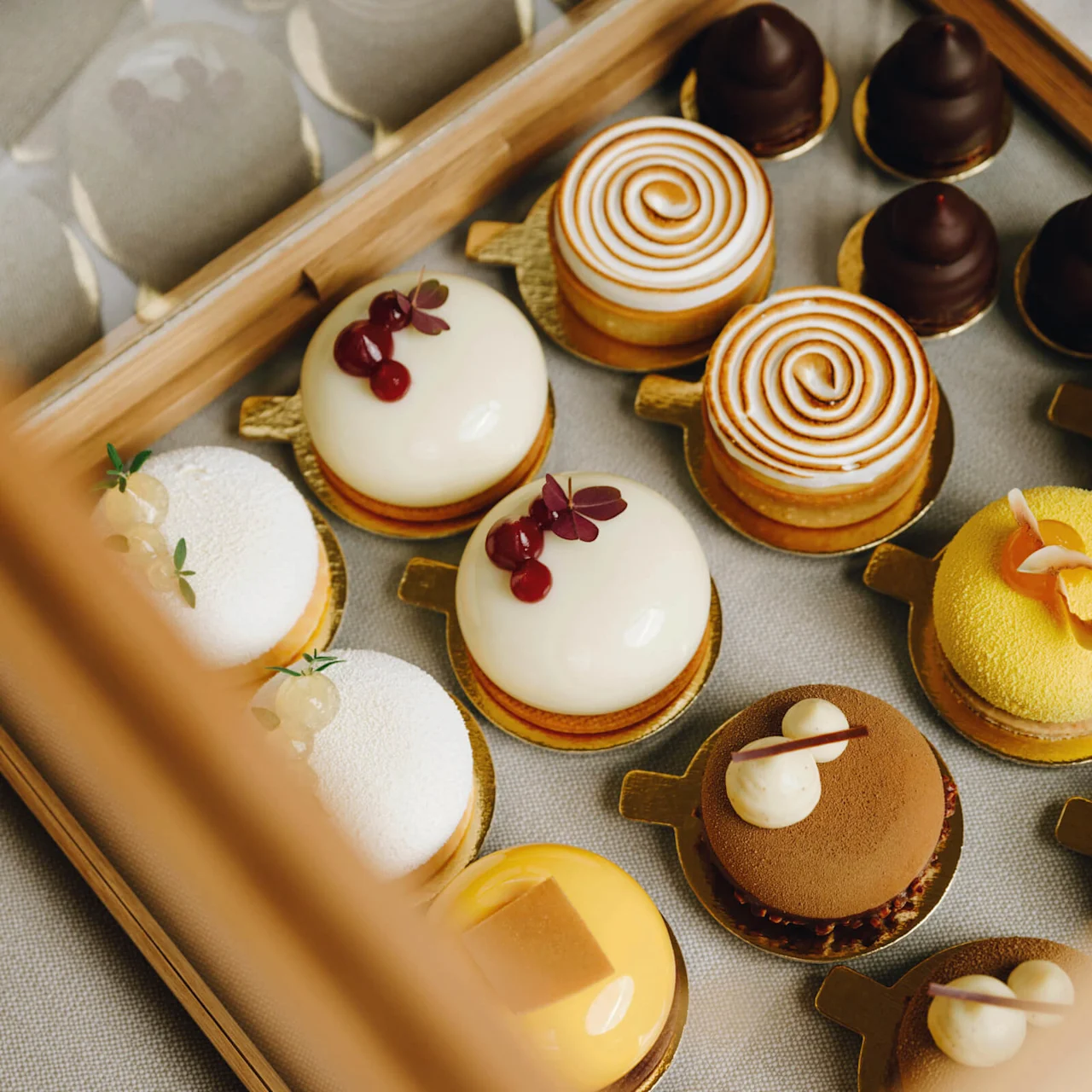 Assorted miniature cakes sit in a wooden box, featuring creamy toppings, swirls, and decorative berries, creating an elegant and colorful presentation against a neutral background.