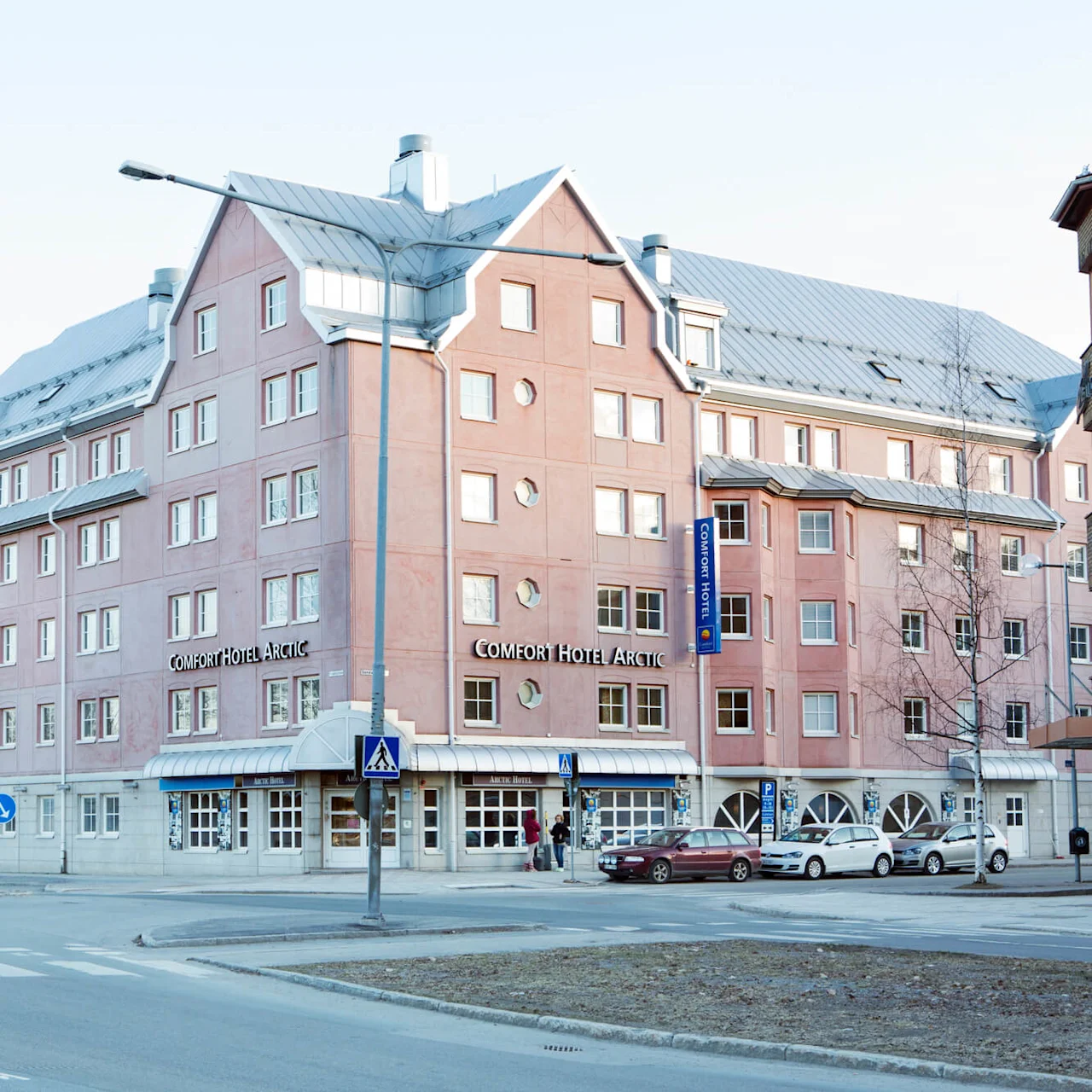 Salmon-colored hotel building with a clear blue sign reading "Comfort Hotel Arctic," surrounded by a street with parked cars and bare trees.