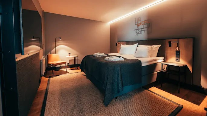 En pent oppredd seng med mørke tepper står i et moderne hotellrom. Myk belysning fremhever en lenestol, sidebord og abstrakt veggkunst. Et teksturert teppe dekker tregulvet.