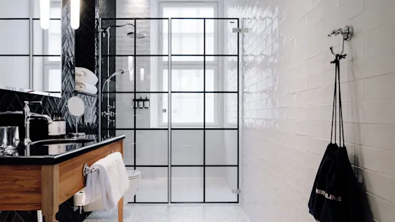 A bathroom features a wooden vanity with a towel, next to a glass shower enclosure. White and black tiles adorn the walls. A bag with "MERLIN" hangs nearby.