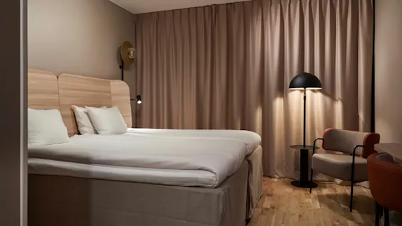 Ett inbjudande hotellrum med en prydligt bäddad säng, varm belysning från en golvlampa och en bekväm fåtölj.