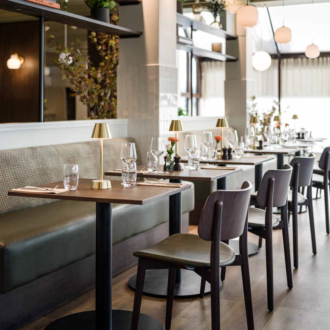 A series of wooden tables with chairs are set for dining, featuring glasses and small lamps, in a modern restaurant with large windows and plants for decoration.