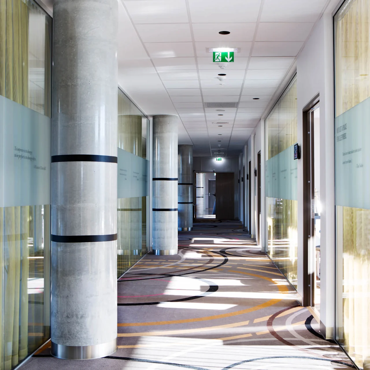 A modern corridor with glass walls features patterned carpet and concrete columns. Text on the glass includes: "NOT JUST A PLACE FOR SLEEPOVERS... Work until your idols become your rivals" and more quotes. 