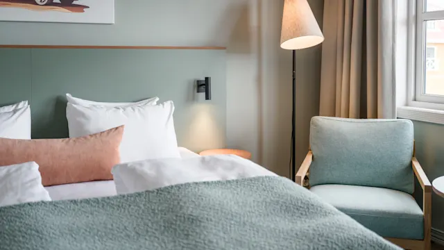 En pent oppredd seng med hvitt og pastellfarget sengetøy står i et koselig rom. Et maleri av en varebil henger over; en gulvlampe og lenestol står ved siden av et vindu.