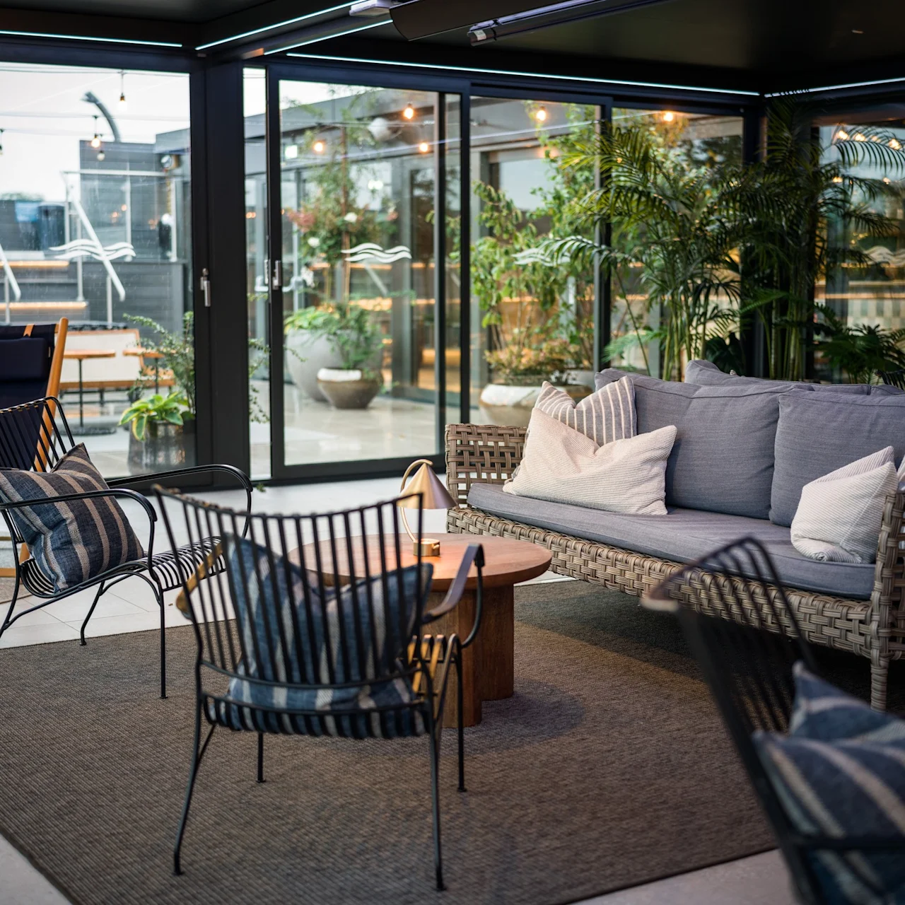 Flätad soffa med kuddar står i en modern inomhuslounge. Svarta metallstolar omger ett träbord, med grönska och glasdörrar som leder till en utomhusaltan.