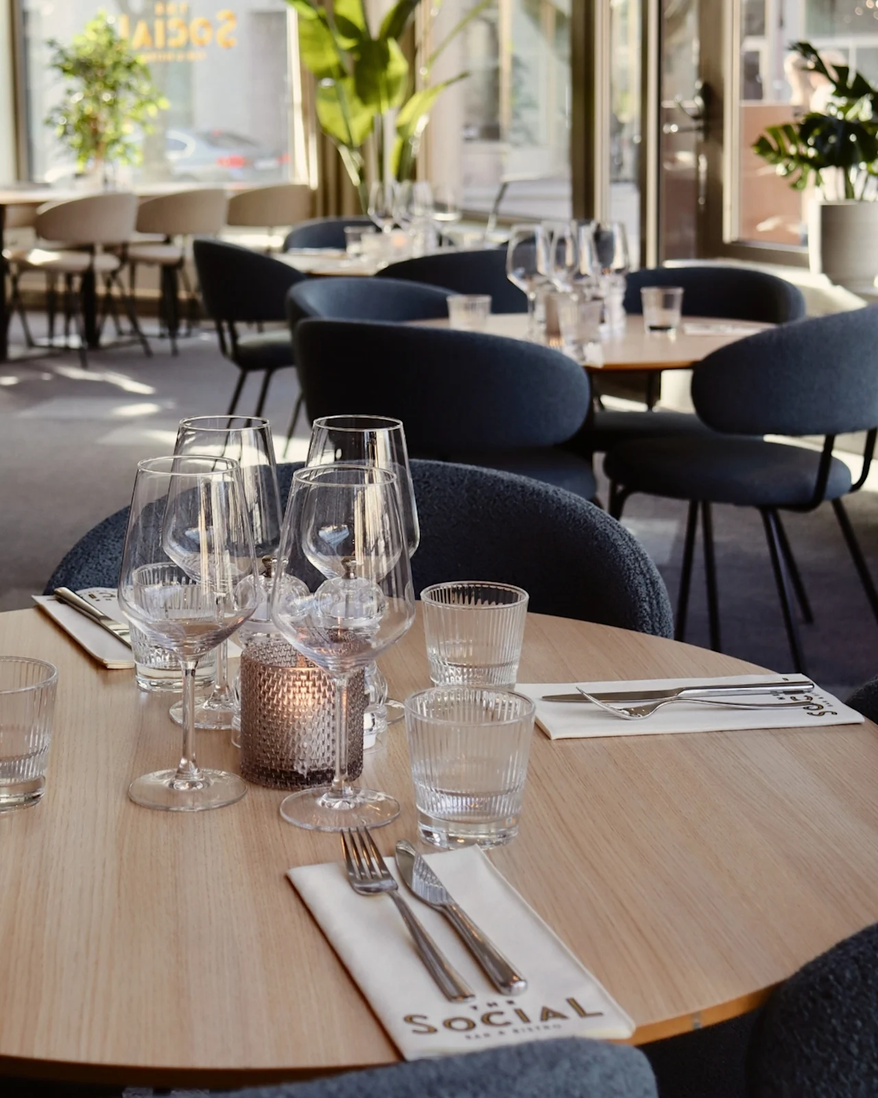 A round wooden table is set with wine glasses and cutlery on napkins labeled "SOCIAL." Chairs surround the table in a bright, plant-filled dining area.