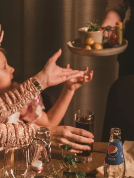 Ett barn och en vuxen sträcker sig mot en serveringsperson som håller en bricka med matvaror. Bordet har ett glas cola, en Pepsi-flaska och dekorativa växter under varm belysning.